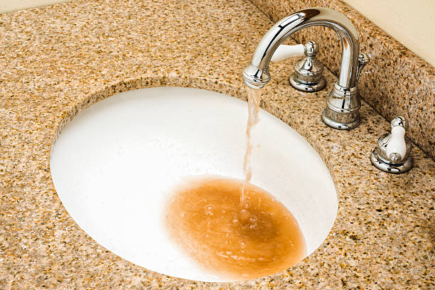 Looking down on brown contaminated dirty water pouring out of a chrome goose neck faucet into a white porcelain sink bowl in a granite vanity top